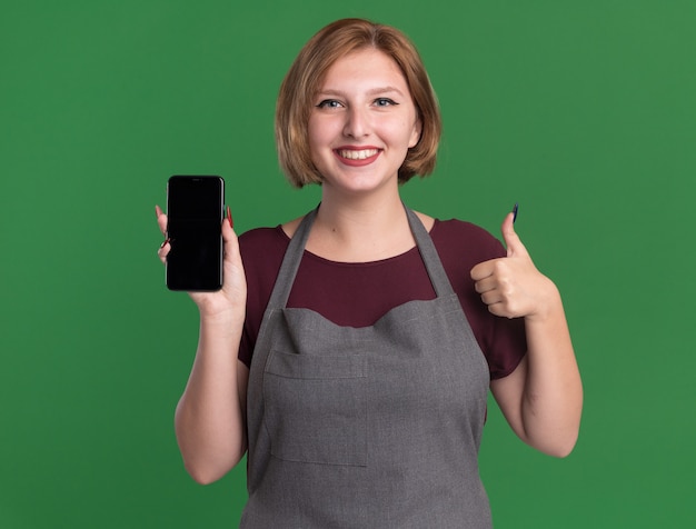 Jonge mooie vrouwenkapper in de smartphone die van de schortholding voorzijde glimlachen die vrolijk tonen die duimen tonen die zich over groene muur bevinden