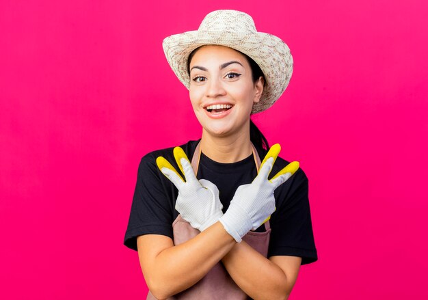 Jonge mooie vrouwentuinman die in rubberhandschoenenschort en hoed het tonen v-teken glimlachen