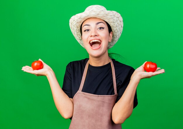 Jonge, mooie vrouwentuinman in schort en hoed die tomaten vasthoudt die glimlachen met een blij gezicht dat over een groene achtergrond staat