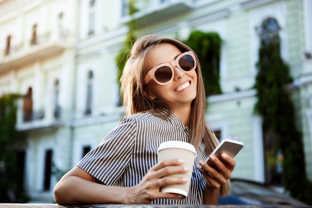 Jonge mooie vrouwenzitting op bank, die koffie en telefoon houdt.