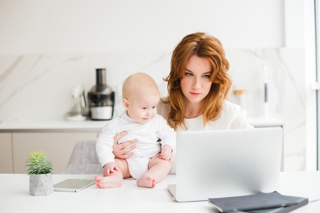 Jonge, mooie zakenvrouw die aan tafel zit en op laptop werkt terwijl ze haar schattige kleine baby vasthoudt