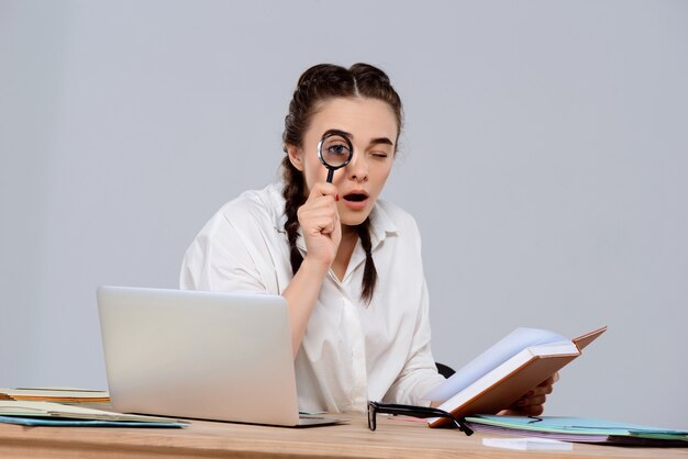 Jonge mooie zakenvrouw zittend op de werkplek, met boek en vergrootglas