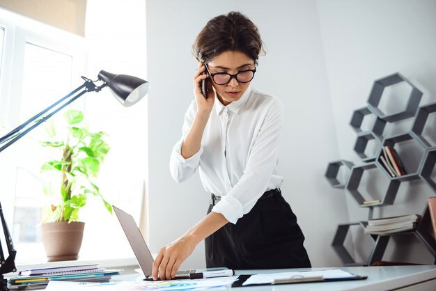 Jonge mooie zekere onderneemster die op telefoon op werkplaats in bureau spreekt.