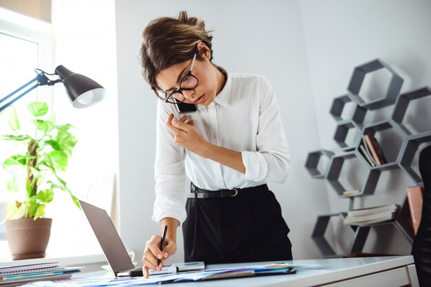 Jonge mooie zekere onderneemster die op telefoon op werkplaats in bureau spreekt.