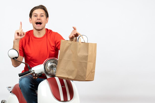 Jonge nieuwsgierige koerierskerel in rood uniform zittend op scooter met papieren zak omhoog op witte muur