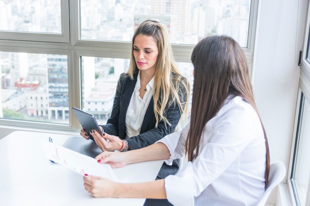 Jonge onderneemster die digitale tabletzitting met haar vrouwelijke collega in het bureau bekijkt