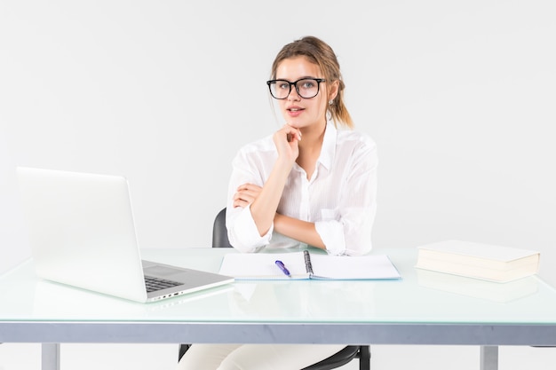 Jonge onderneemsterzitting bij een bureaulijst met laptop, die de camera bekijkt die op witte achtergrond wordt geïsoleerd