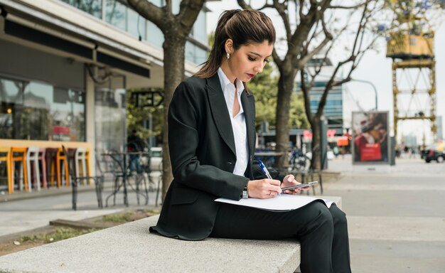 Jonge onderneemsterzitting op bank die op papier schrijven die mobiele telefoon in hand houden