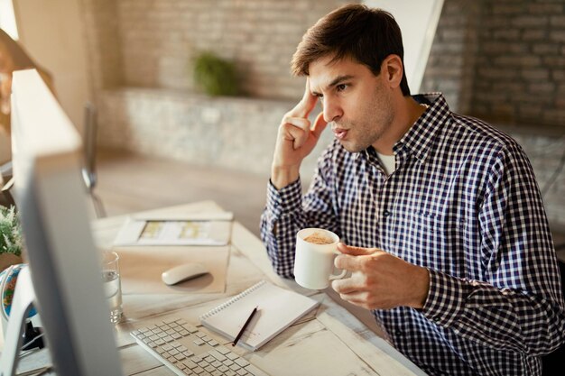 Jonge ondernemer voelt zich bezorgd terwijl hij op een computer werkt en koffie drinkt op kantoor
