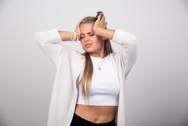 Jonge onrustige vrouw in witte uitrusting die zich op grijze achtergrond bevindt.