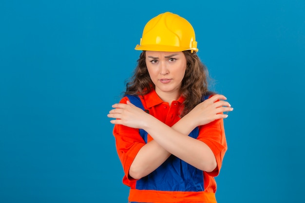 Jonge ontevreden bouwersvrouw in eenvormige bouw en veiligheidshelm die zich met gekruiste wapens op borst over geïsoleerde blauwe achtergrond bevinden
