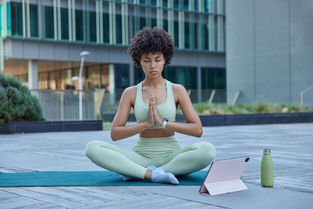 Jonge ontspannen vrouw doet yoga op mat zit in lotushouding houdt handpalmen tegen elkaar gedrukt ogen dicht ademt diep kijkt instructievideo