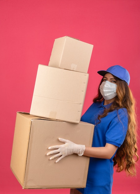 Jonge over gespannen leveringsvrouw met krullend haar die blauw poloshirt en pet in medisch beschermend masker dragen die zich met dozen over geïsoleerde roze achtergrond bevinden