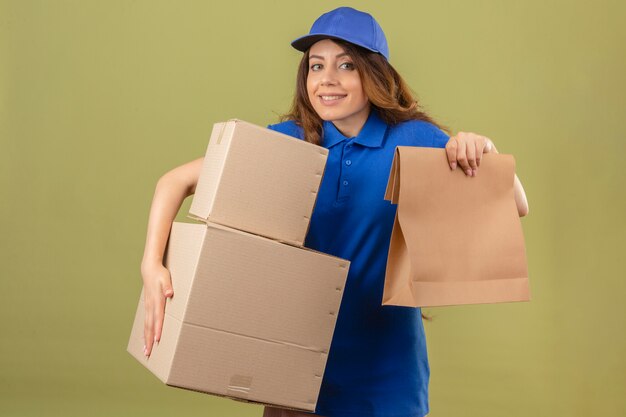 Jonge over gespannen leveringsvrouw met sluwe blik die blauw poloshirt en GLB draagt die zich met stapel pakketten over geïsoleerde groene achtergrond bevinden