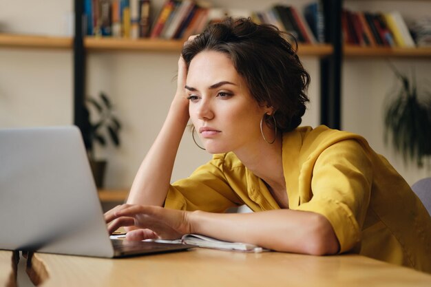 Jonge, overstuur vrouw die aan het bureau zit en vermoeid werkt aan een nieuw project met een laptop in een modern kantoor
