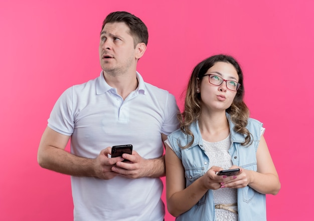 Jonge paarman en vrouw die in vrijetijdskleding smartphones houden die opzij kijken met peinzende uitdrukking die zich over roze muur bevindt
