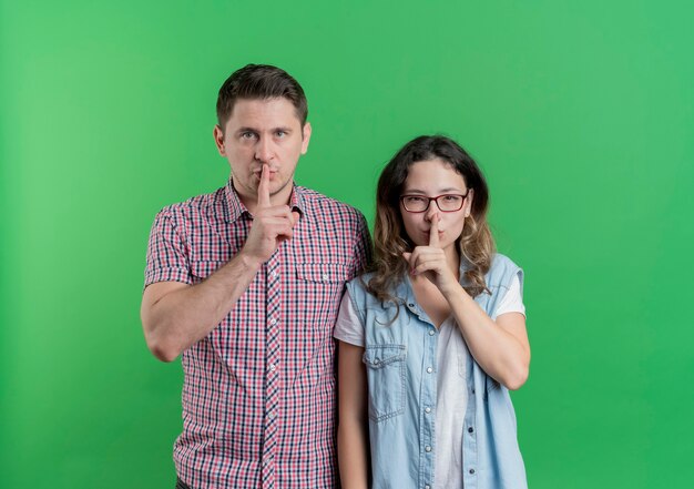 Jonge paarman en vrouw die in vrijetijdskleding stilte gebaar met vinger op lippen maken die zich over groene muur bevinden