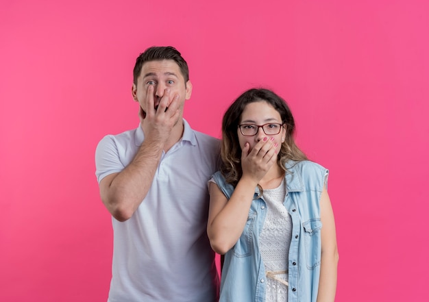 Jonge paarman en vrouw in vrijetijdskleding die mond behandelen met handen die worden geschokt status over roze muur