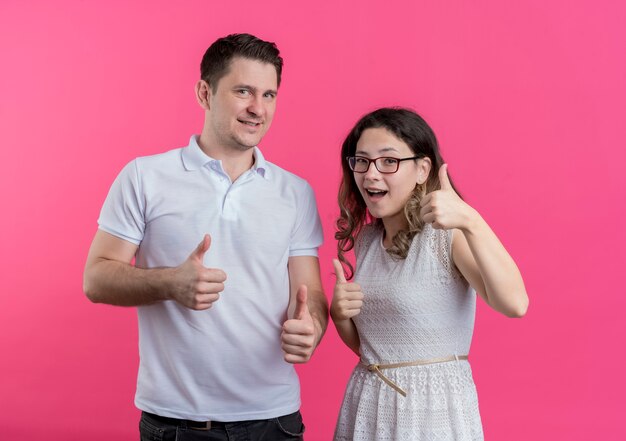 Jonge paarman en vrouw in vrijetijdskleding die vrolijk glimlachen tonen duimen omhoog die zich over roze muur bevinden