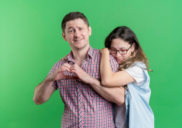 Jonge paarman en vrouw in vrijetijdskleding die zich gelukkig man bevinden die hartgebaar over groene muur tonen