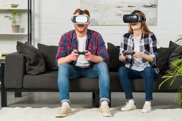 Jonge paarzitting op de bank die virtuele werkelijkheidsglazen gebruiken die het videospelletje met bedieningshendel spelen