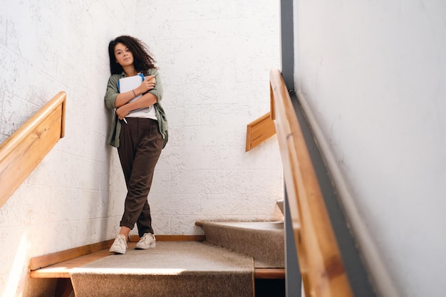 Jonge peinzende student meisje met donker krullend haar in kaki shirt en broek staande op trappen in universiteit met notitieblok en kopje koffie om in handen te gaan en zorgvuldig in de camera kijken