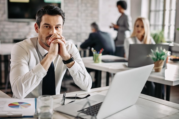 Jonge peinzende zakenman die aan het bureau op kantoor werkt en aan iets denkt Er zijn mensen op de achtergrond