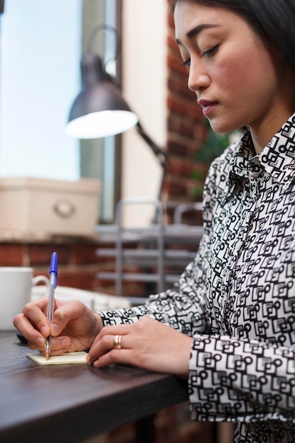 Jonge productieve aziatische zakenvrouw die belangrijke aantekeningen maakt terwijl ze aan het bureau zit in de werkruimte van het bedrijf. werknemer van het marketingbureau die persoonlijke informatie van de klant met betrekking tot samenwerking opschrijft.