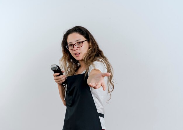 Jonge professionele kapper vrouw in schort bedrijf trimmer maken kom hier gebaar met hand glimlachen kijken voorzijde staande over witte muur