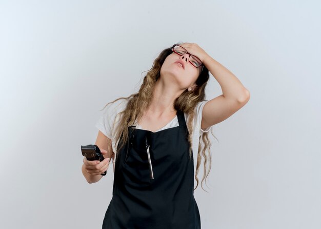 Jonge professionele kapper vrouw in schort met trimmer met haar hand op voorhoofd wordt moe en verveeld staande over witte muur