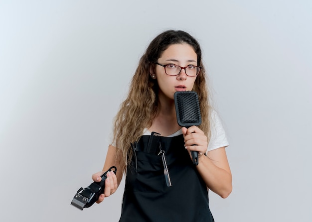 Jonge professionele kappervrouw die in de trimmer en de haarborstel van de schortholding voorzijde met ernstig gezicht bekijken die zich over witte muur bevinden