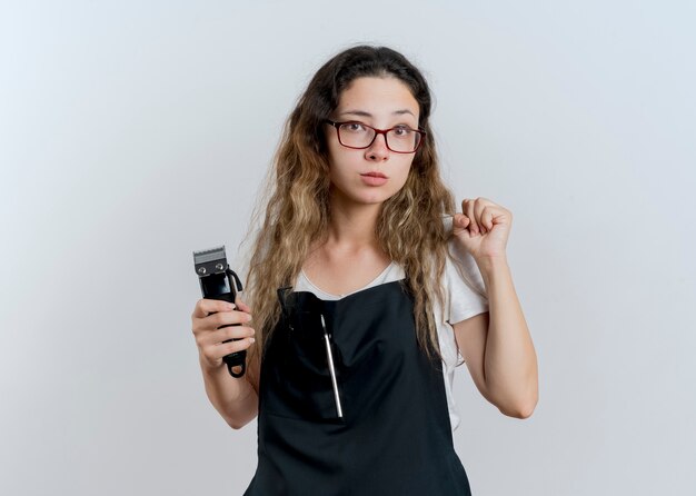 Jonge professionele kappervrouw die in de trimmer van de schortholding voorzijde met ernstig gezicht bekijkt die vuist zich over witte muur balt