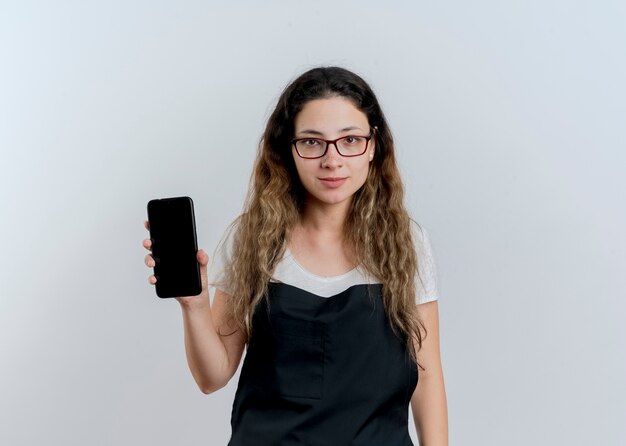 Jonge professionele kappervrouw die in schort smartphone tonen die voorzijde glimlachend zelfverzekerd over witte muur bekijken kijken