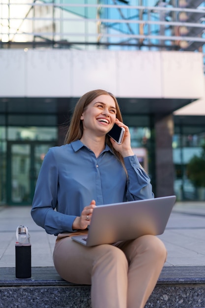 Jonge professionele vrouw zittend op de trap voor glazen gebouw, laptop op schoot houden en praten op mobiele telefoon