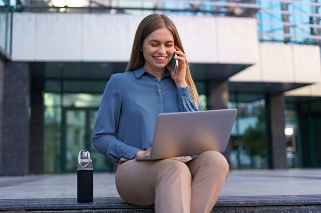 Jonge professionele vrouw zittend op de trap voor glazen gebouw, laptop op schoot houden en praten op mobiele telefoon