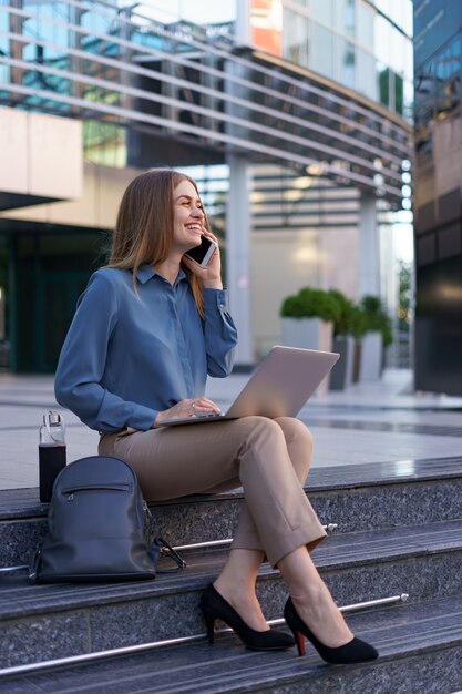 Jonge professionele vrouw zittend op de trap voor glazen gebouw, laptop op schoot houden en praten op mobiele telefoon