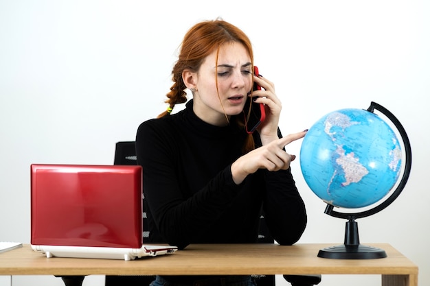 Jonge reisagent vrouw zit achter bureau met laptopcomputer en geografische wereldbol praten op een mobiele telefoon.