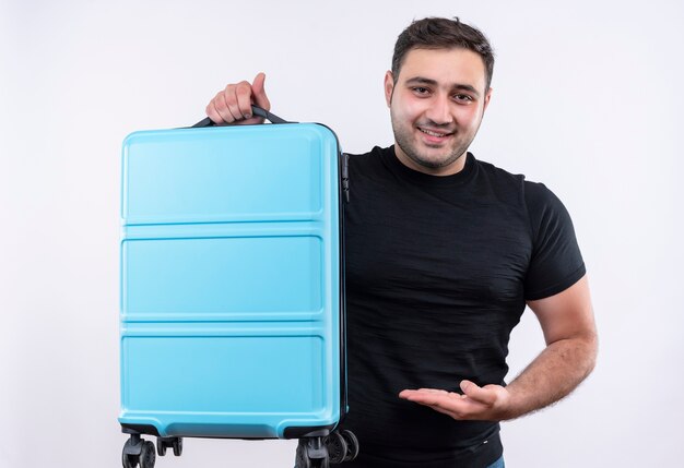 Jonge reiziger man in zwarte t-shirt bedrijf koffer presenteert het met arm glimlachend zelfverzekerd staande over witte muur