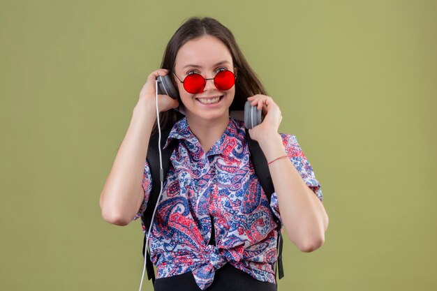 Jonge reiziger vrouw dragen rode zonnebril en met rugzak luisteren naar muziek met behulp van koptelefoon lachend met blij gezicht staande over groene achtergrond