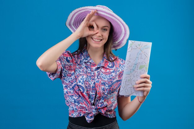 Jonge reiziger vrouw in zomer hoed bedrijf kaart positief en gelukkig doen ok teken kijkt door dit bord, lacht vrolijk over blauwe muur