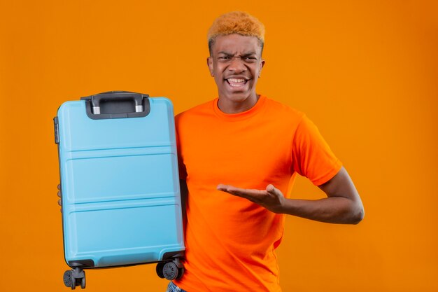 Jonge reizigersjongen die een oranje t-shirt draagt en een koffer vasthoudt die het presenteert met de arm van zijn hand, ontevreden gevoel geïrriteerd staande over oranje muur