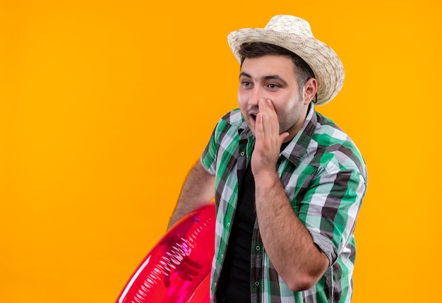 Gratis foto jonge reizigersmens in geruit overhemd en zomerhoed die opblaasbare ring houdt die een geheim vertelt met hand dichtbij mond die zich over oranje muur bevindt