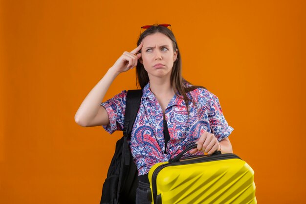 Jonge reizigersvrouw die een rode zonnebril op het hoofd draagt en met rugzak staat met koffer wijzend tempel met fronsend gezicht herinnert zich dat ze het belangrijke niet vergeet dat over oran stond