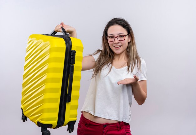 Jonge reizigersvrouw die in de witte koffer van de t-shirtholding het voorstellen met wapen van haar hand die zich over witte muur bevinden