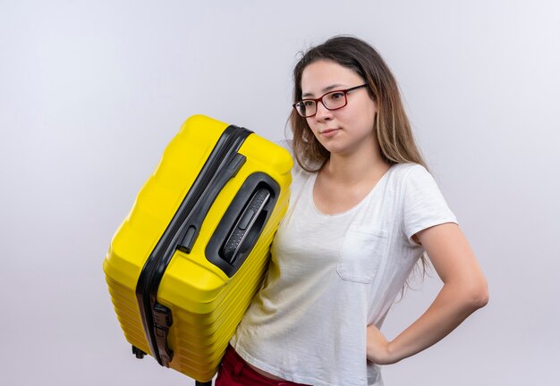 Jonge reizigersvrouw die in de witte koffer van de t-shirtholding opzij verbaasd status over witte muur kijken