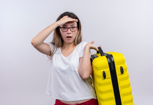 Jonge reizigersvrouw die in de witte koffer van de t-shirtholding verbaasd en verbaasd over witte muur kijken