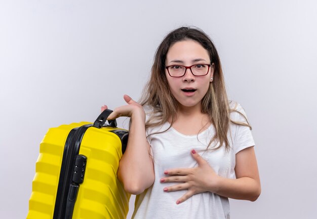 Jonge reizigersvrouw die in de witte koffer van de t-shirtholding verbaasd en verbaasd over witte muur kijken