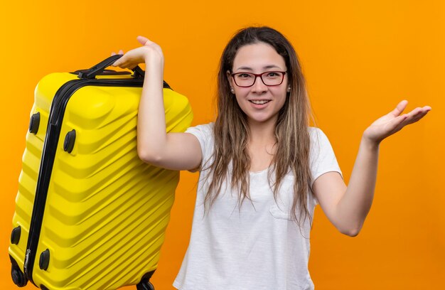 Jonge reizigersvrouw die in de witte koffer van de t-shirtholding verward kijken hebbend twijfels glimlachend status over oranje muur