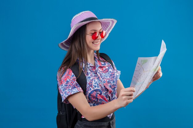 Jonge reizigersvrouw die in de zomerhoed rode zonnebril dragen die kaart houden die het bekijken glimlachend met gelukkig gezicht over blauwe muur