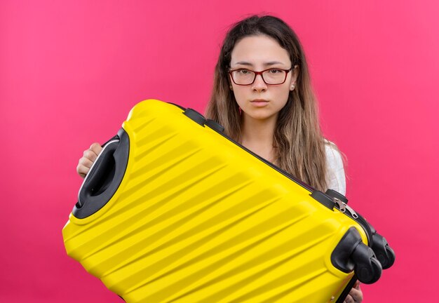 Jonge reizigersvrouw die in witte de reiskoffer van de t-shirtholding met ernstige uitdrukking kijken die zich over roze muur bevindt